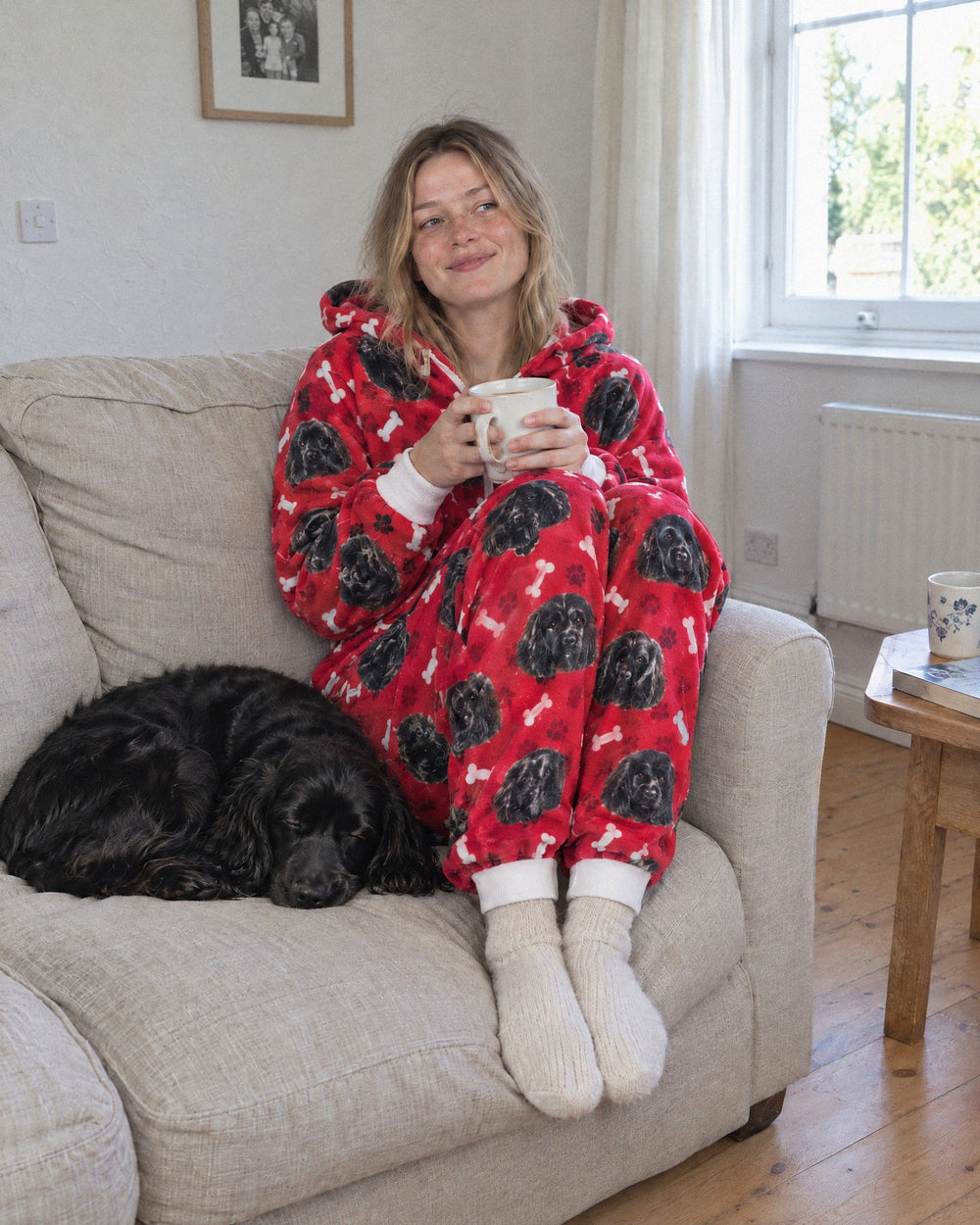 Woman in a red personalised onesie printed with her black spaniel, holding a mug of tea while the spaniel sleeps beside her