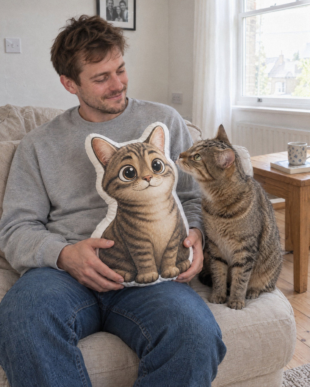 Man holding a cartoon cushion of his tabby cat while the real cat sniffs it on the sofa
